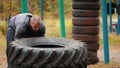Tough tattooed man bodybuilder in gloves pushes over the truck tire on the ground - training in the autumn forest