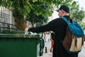 A tourist with a backpack and in a baseball cap on the street throws a plastic bottle into a container with waste. Care