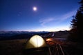 Tourist camping near forest in the mountains. Illuminated tent and campfire under evening sky full of stars and the moon Royalty Free Stock Photo