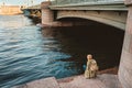 A tourist girl with a backpack rests on the river embankment against the background of the bridge and looks at the city.