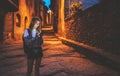 Tourist girl blonde with glasses and camera using mobile phone traveling in night old historical city Europe, young hipster plans