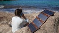 A tourist girl is talking on a smartphone on the seashore. The phone is charged by a solar panel that lies on a stone Royalty Free Stock Photo