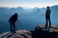 Tourist guide and photo enthusiast stay with tripod on cliff and thinking. Dreamy fogy landscape, blue misty sunrise in a beautifu Royalty Free Stock Photo