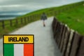 Tourist holding badge with sign Ireland and Irish flag in focus, tourist walking on path to Cliff of Moher out of focus.