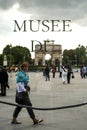 Tourist passing by behind the logo of Louvre Museum Musee du Louvre, in front of Jardin des Tuileries Garden.