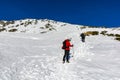 Tourist in a red jacket in a helmet with a camera and trekking poles following a group of people on a steeply snowy slope
