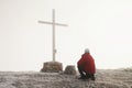Tourist in red is kneeling at cross memorial on mountain peak. Man is watching into misty Alpine valley bellow.