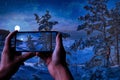 Tourist taking photo of magic winter night in a snowy sea coast surrounded by covered in snow fir trees with full moon in a starry Royalty Free Stock Photo