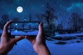 Tourist taking photo of magic winter night with starry sky and full moon in snowy park with beautiful bridge over small pond, Royalty Free Stock Photo