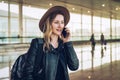 Tourist woman in hat and with backpack is standing at airport and talking on cell phone.Girl stands, uses digital gadget Royalty Free Stock Photo