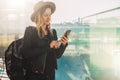 Tourist woman in hat, with backpack stands at airport, uses smartphone. Girl checks email, chatting, browsing internet. Royalty Free Stock Photo