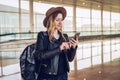 Tourist woman in hat, with backpack stands at airport, uses smartphone. Girl checks email, chatting, browsing internet. Royalty Free Stock Photo