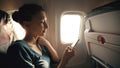 Tourist woman sitting near airplane window at sunset and using mobile phone during flight Royalty Free Stock Photo