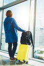 Tourist with yellow suitcase backpack is standing at airport on background large window, traveler girl waiting in departure lounge Royalty Free Stock Photo