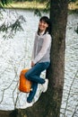 Tourist young smiling girl with orange backpack in hands standing near to big tree on bank of mountain lake surrounded by forest Royalty Free Stock Photo