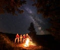 Tourists sit on log by fire under fir trees on the background of sky strewn with stars