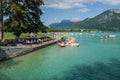 A sunny view on Lake Annecy, France, with pedal boats on the quay and tourists in the park