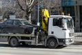 A tow truck with hook and chain transports a car without a front wheel along the city street during the day
