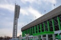 A tower with floodlights illuminating a sports stadium in the daytime against a cloudy sky and part of the grandstand