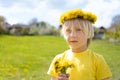 Towheaded boy in wreath of flowers with bouquet of dandelions stand in meadow. Summer vacation time