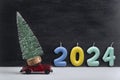 Toy car carries miniature Christmas tree and colored numbers 2024, New Year. Delivery of Christmas trees concept