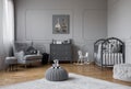 Toy on grey pouf in the middle of trendy baby bedroom with grey wooden cradle, chest of drawers and comfortable armchair, real