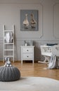 Toy on grey pouf in trendy baby bedroom with cradle and white wooden furniture, real photo with poster on the wall