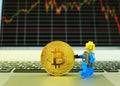 A toy man carrying a gold bitcoin on a notebook computer with stock graph in the background