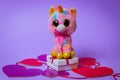 Toy Pink Unicorn sits on a white gift box with a Red Ribbon on paper hearts on a purple background Royalty Free Stock Photo