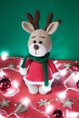 Toy small Christmas deer on a red and green background with decorations and lights from a garland. Cute handmade knitted deer in a