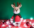Toy small Christmas deer on a red and green background with decorations and lights from a garland. Cute handmade knitted deer in a