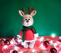 Toy small Christmas deer on a red and green background with decorations and lights from a garland. Cute handmade knitted deer in a