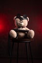 a toy teddy bear with a whip, dressed in leather belts and a mask, an accessory for BDSM games on a dark red texture