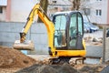 Tracked orange mini excavator digger near road with green trees and concrete fence on background with copyspace. Royalty Free Stock Photo
