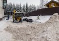 A tractor with a bucket rakes snow into a pile in the yard of the house.