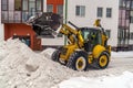 A tractor with a bucket rakes snow into a pile in the yard of the house.