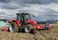 Tractor plows the field in front of some agricultural buildings on a sunny spring day