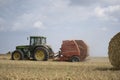 A tractor uses a trailed bale machine to collect straw in the field and make round large bales.