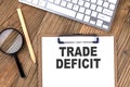 TRADE DEFICIT text on paper clipboard with magnifier and keyboard on wooden background