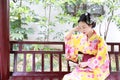 Traditional Asian Japanese beautiful Geisha woman wears kimono hold a fan sit on a bench in a summer nature garden