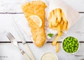 Traditional British Fish and Chips with tartar sauce on chopping board with fork and knife and green peas on white wooden