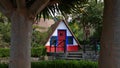 Traditional Madeiran house in triangle shape with red, white and blue facade viewed through the trunks of tropical trees.