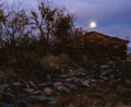 A traditional stone hut, locally known as a Channi, set against the backdrop of a rising moon and the majestic Himalayas.