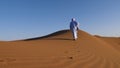 A traditionally dressed Moroccan man wearing a blue gandoura, djellaba and a turban walks on a sand dune, Erg Chegaga desert, Moro