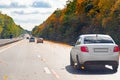 Traffic of cars by day along a busy road among autumn trees and flying yellow leaves.