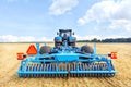 A trailed blue harrow on an agricultural tractor against the backdrop of a harvested yellow wheat field on a warm fall day.