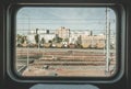 Train window view with old railway, clear blue sky, white and red brick buildings.