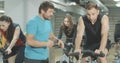 Trainer in blue T-shirt endorsing young handsome sportsman cycling in gym. Professional employee working in sports club