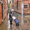 Trandy fashionable mature woman with gaiters, boots and suitcase at high tide in old narrow flooded street Royalty Free Stock Photo