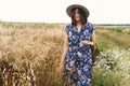 Tranquil summer in countryside. Stylish young woman in blue vintage dress and hat walking with white wildflowers in straw basket